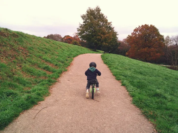 A child riding a bicycle up a red path between two patches of grass with the tops of trees poking out above the brow of the hill