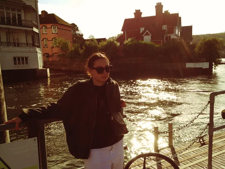 A woman stands on a dockside in front of a body of water and some period buildings bathed in warm golden light
